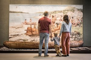 Derrière un canot mi'gmaq posé au sol, une famille admire une fresque représentant un campement mi'gmaq.